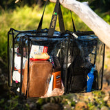 Clear large black trim camping bag with black handles filled with toiletries, hanging on tree outdoors.