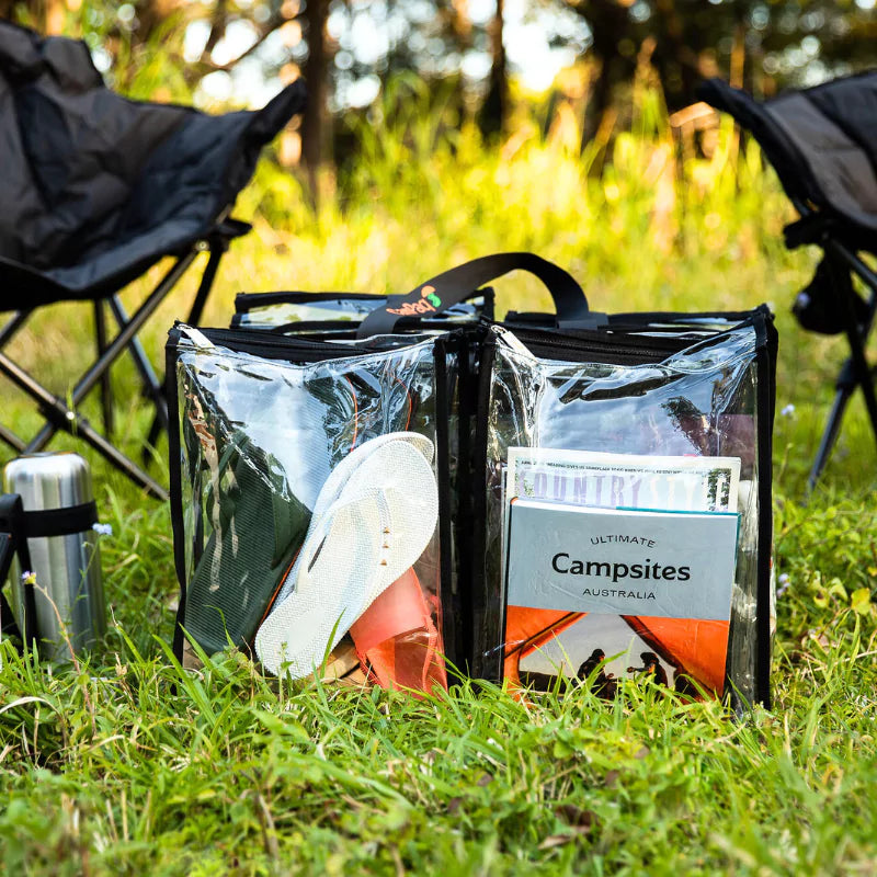 Large black trim clear camping bag filled with books shoes and camp gear on grass outdoors in front of camp chairs