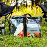 Large black trim clear camping bag filled with books shoes and camp gear on grass outdoors in front of camp chairs