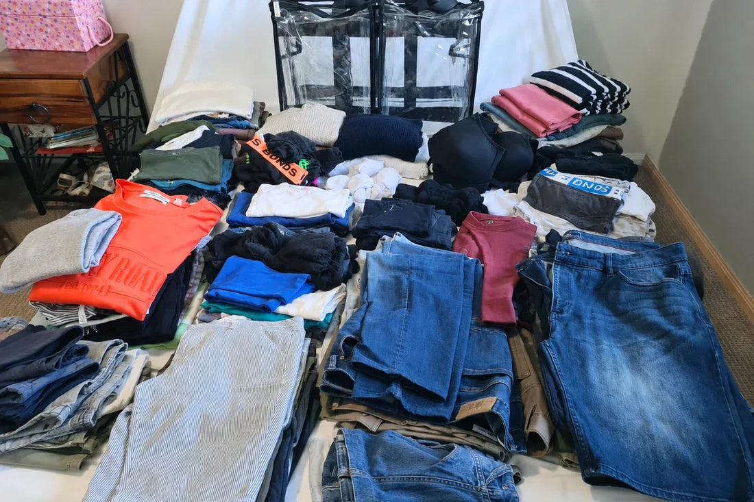 Pile of clothes on bed beside clear camping bag