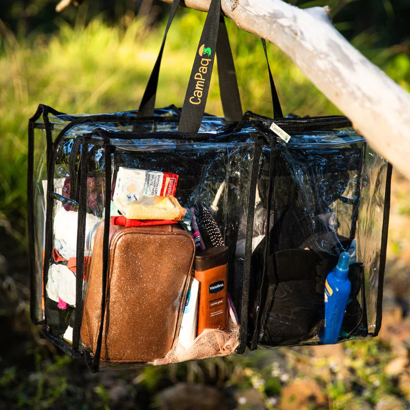 Clear large black trim camping bag with black handles filled with toiletries, hanging  on tree outdoors.