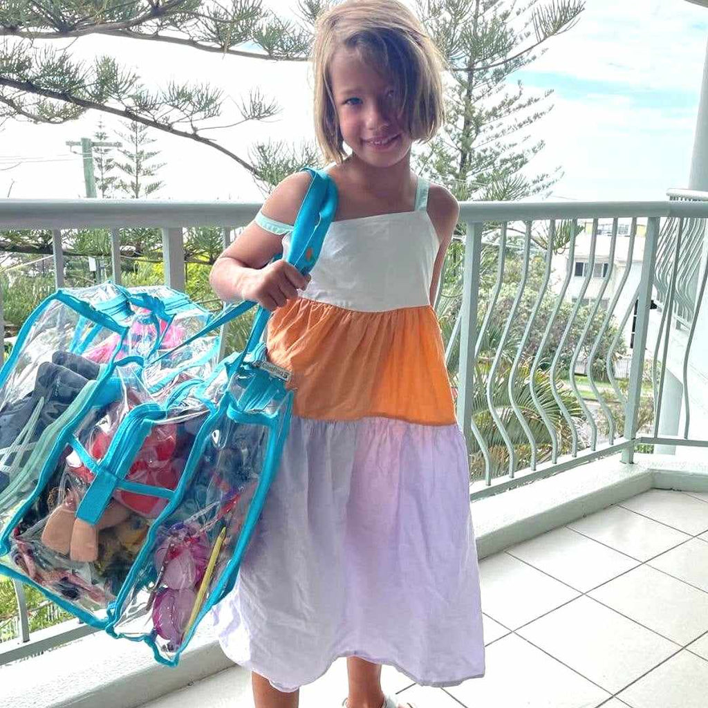 Young girl standing on balcony of holiday unit holding blue medium bag full of beach gear