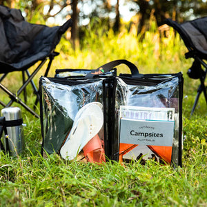 Large black trim clear camping bag filled with books shoes and camp gear on grass outdoors in front of camp chairs