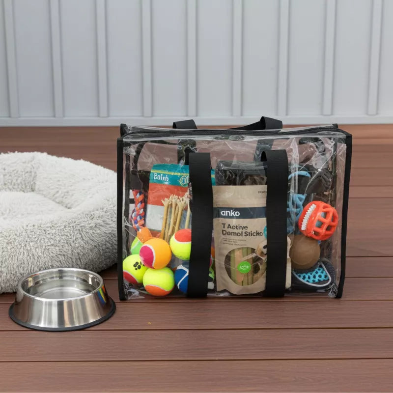 Medium black clear bag filled with dog toys and treats sitting on a wooden deck next to a metal bowl and dog bed