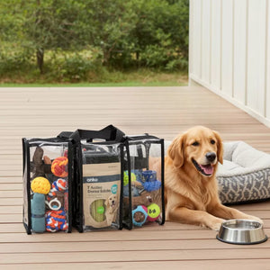 Dog sitting on a wooden deck beside medium black trim clear bag containing dog toys and treats