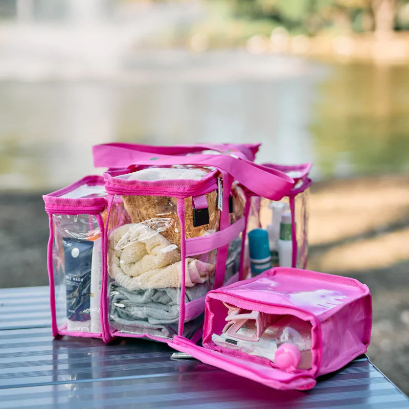Clear hot pink CamPaq toiletry bag filled with toiletries sitting on outside table and showing one detachable opaque pocket with toiletry contents.