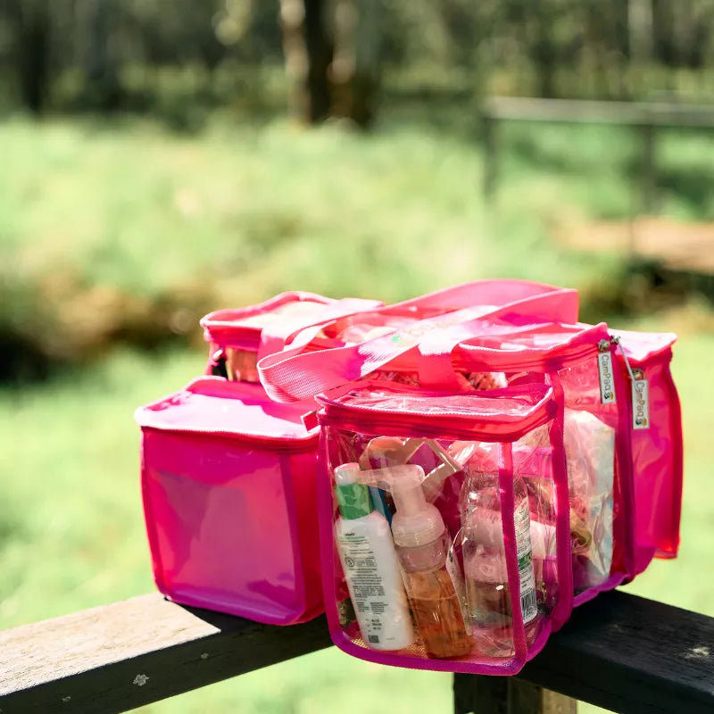 Hot pink clear CamPaq toiletry bag sitting on bench showing opaque pocket and filled with toiletries